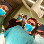 A group of medical professionals wearing scrubs, surgical masks, and caps posing together in an operating room. They appear to be preparing for or concluding a surgical procedure.