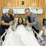 A smiling woman in a hospital bed gives a thumbs up, flanked by two men, one in scrubs and the other in a suit, both also giving thumbs up.